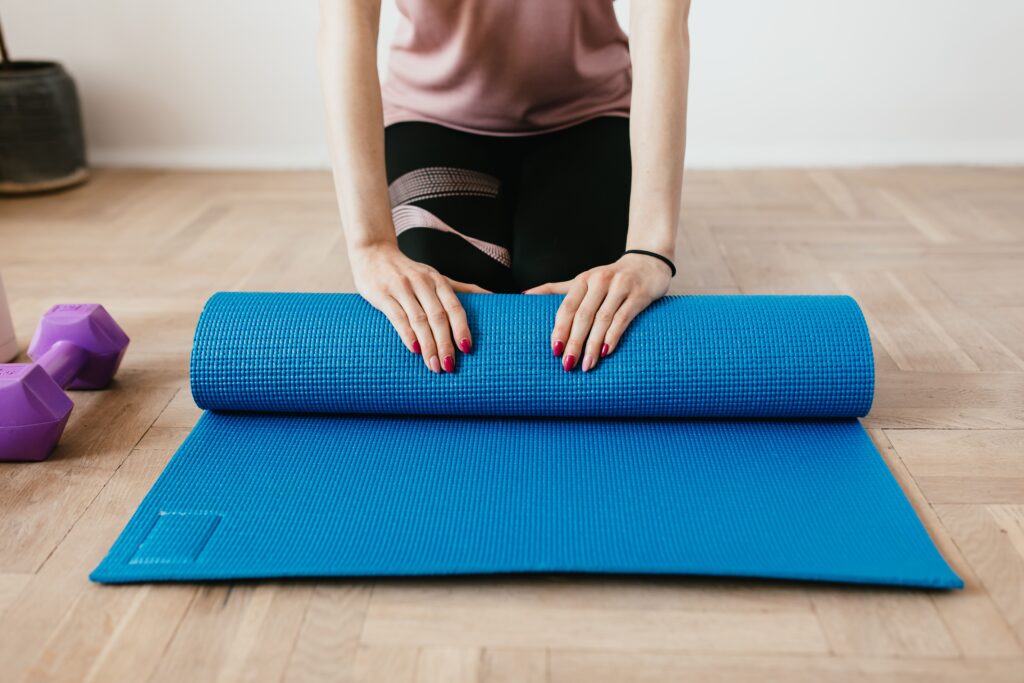 Unrecognizable female athlete in sportswear unfolding blue mat for fitness training on floor near dumbbells at home in daylight